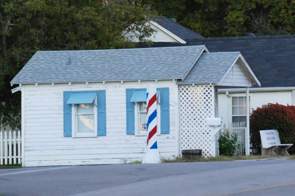 Front view of the small  white wood-sided barber shop.  There is a large barber pole to the left of the building.